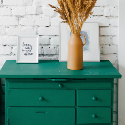 A white brick wall with a short green dresser that has a brown vase and two white picture frames on it.