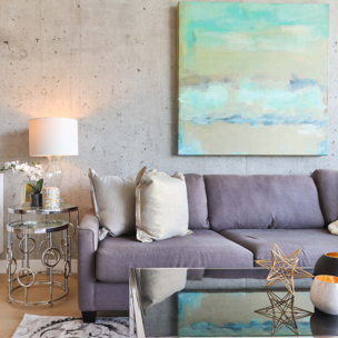 A living room with a light gray couch and a white pillow on it, and a pastel blue painting hanging on the wall above the couch.